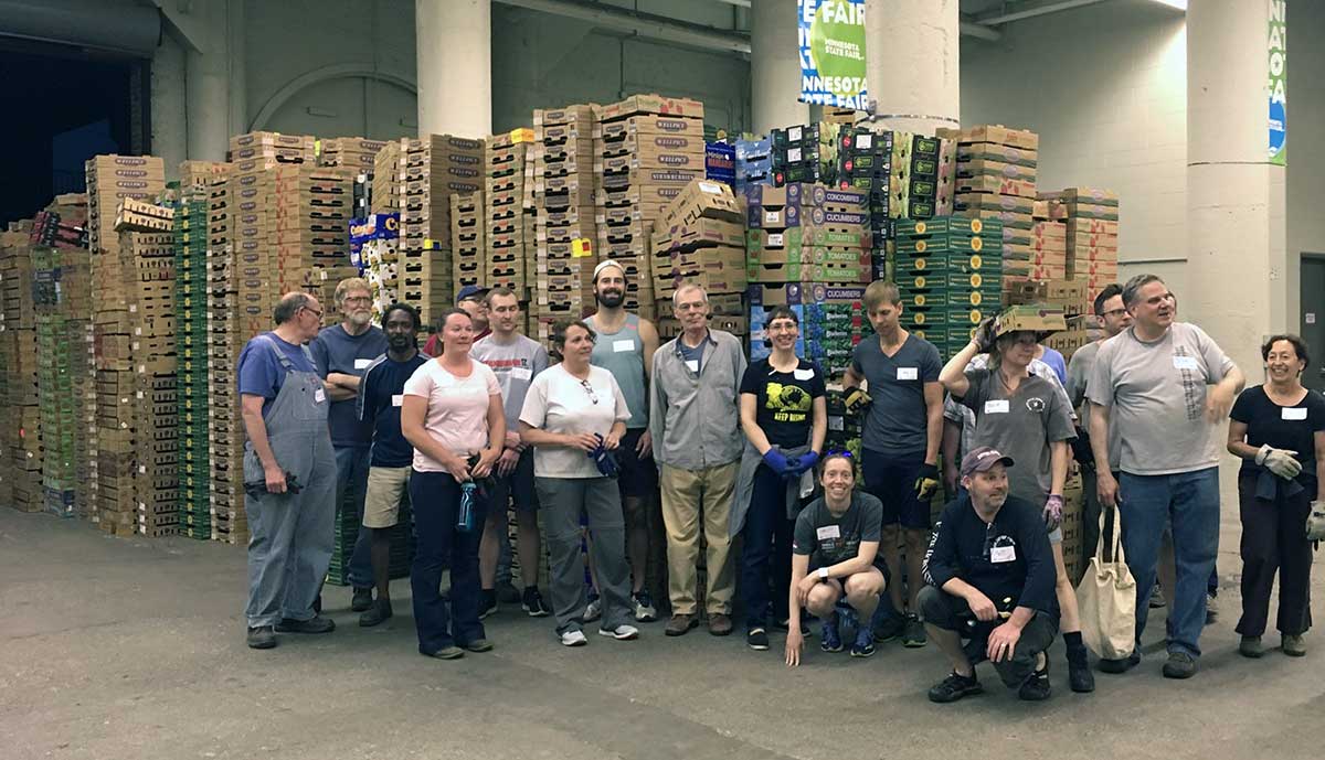 18 people in work clothes standing and squatting in front of piles of shallow cardboard boxes, pillars in the background