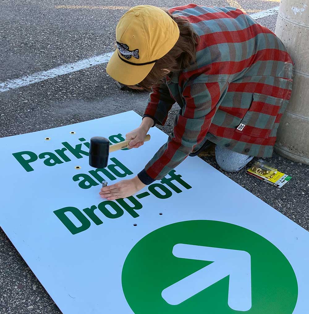 Person kneeling on the ground, using a mallet to put a grommet into a sign that says Parking and Drop-off
