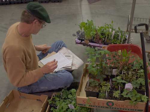 Man with cap sitting on the floor, writing on paper with many plants in boxes and wagon in front of him