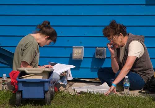 Two women sitting on grass, face to face, in front of a blue wall, each studying a newspaper