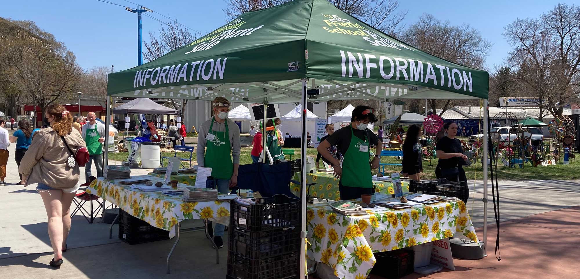 Green popup tent labeled INFORMATION, people with plant sale aprons at tables underneath it