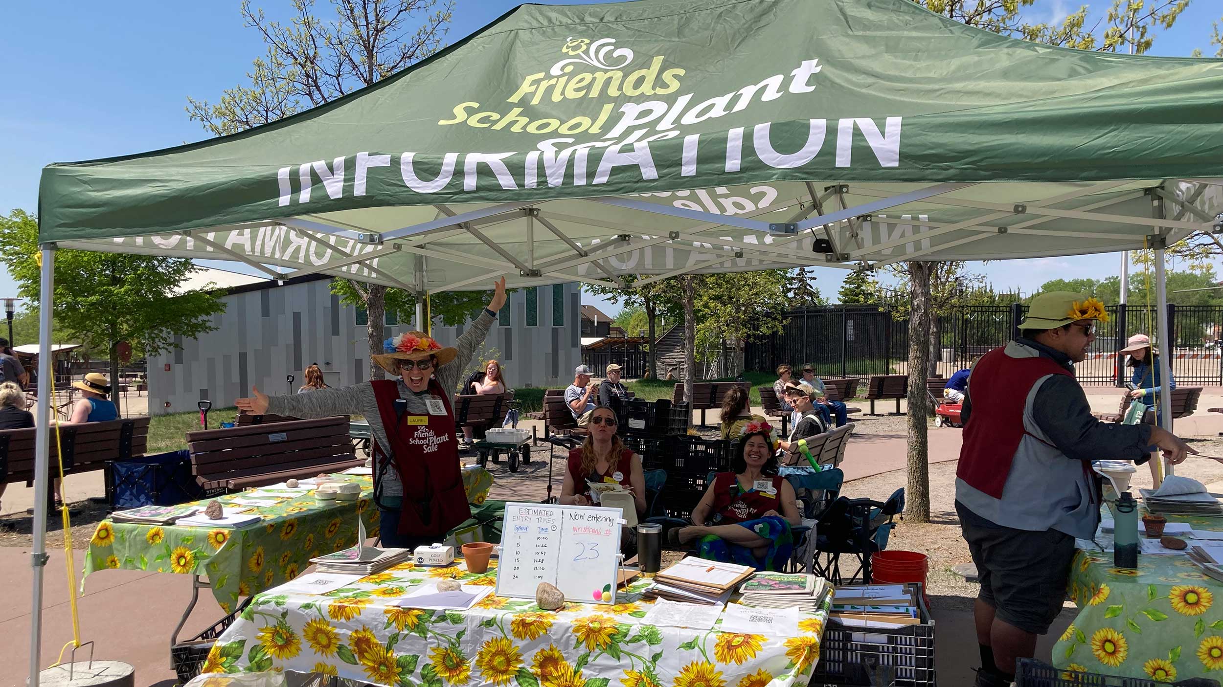 Green popup tent labeled INFORMATION, people with plant sale aprons at tables underneath it