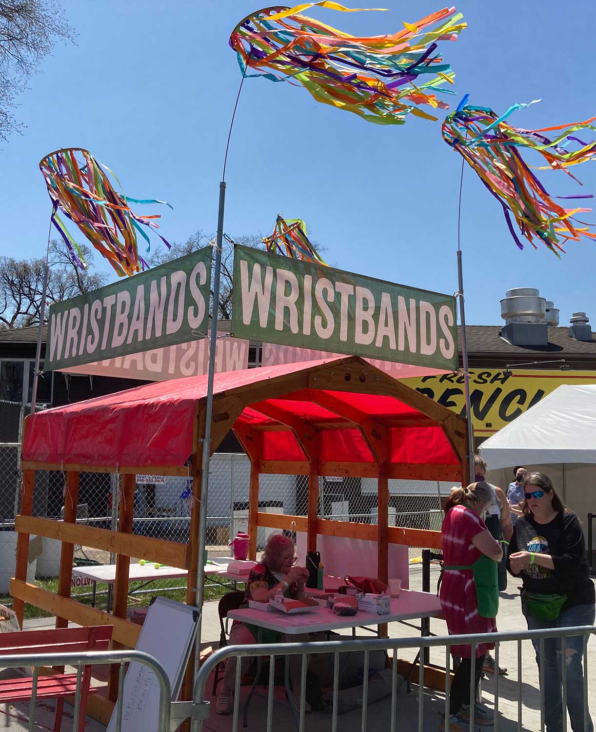 Red-roofed tent with green signs reading WRISTBANDS, colorful streamers flying overhead