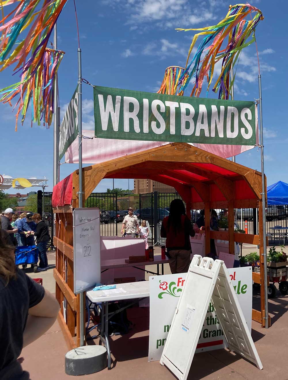 Red-roofed tent with green signs reading WRISTBANDS, colorful streamers flying overhead