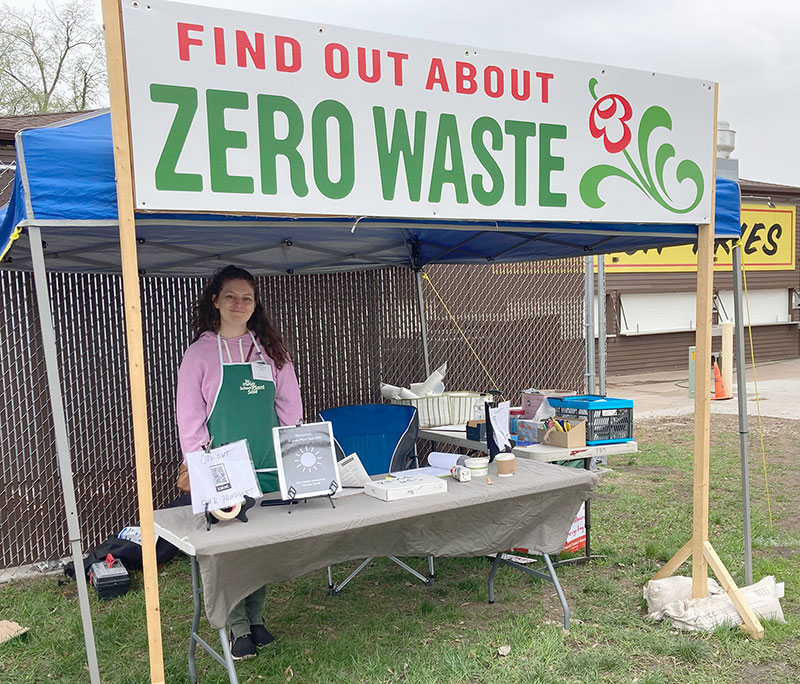 Volunteer standing at a table under a banner that reads Find out about ZERO WASTE