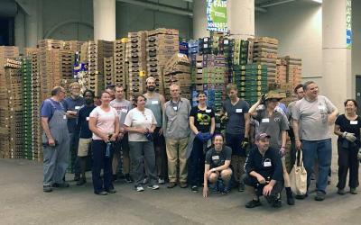 18 people in work clothes standing and squatting in front of piles of shallow cardboard boxes, pillars in the background