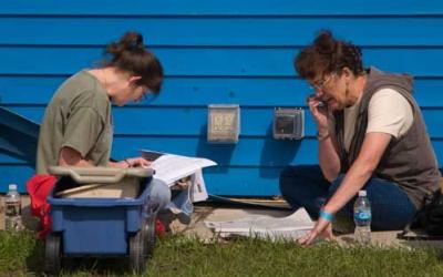 Two women sitting on grass, facing each other, looking at a newspaper